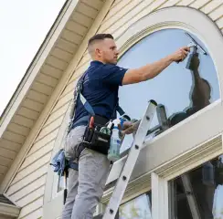 Professional window cleaner on a ladder using a squeegee to clean an exterior arched window, Columbus, GA.
